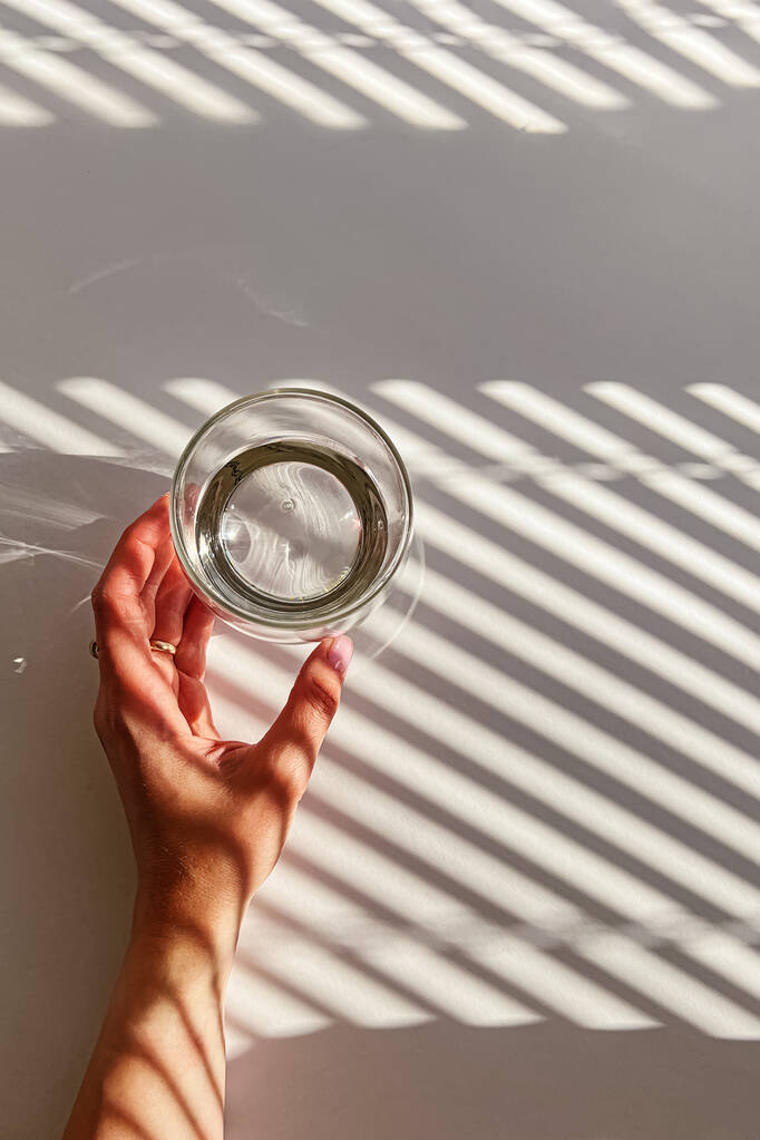 Γυναικείο χέρι αγγίζει ένα ποτήρι καθαρό νερό, θέα από την κορυφή, λευκό τραπέζι, αφηρημένες σκιές, φως το πρωί - Φωτογραφία, εικόνα