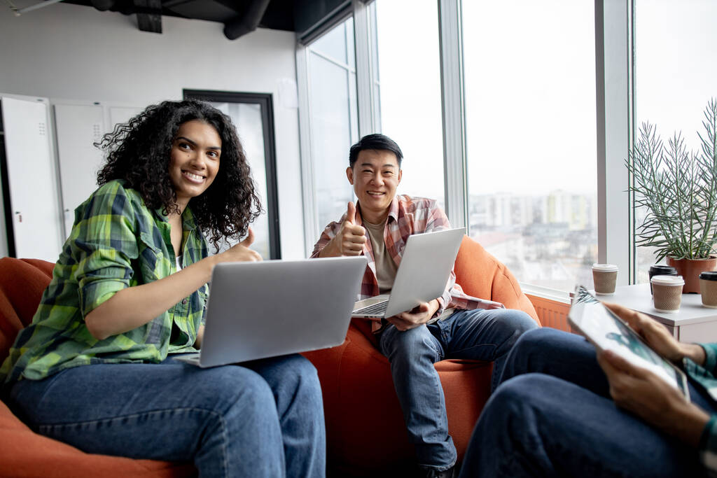 Attractive african american lady and asian man in everyday wear looking at computer screen while sitting on soft seats in office showing thumb up. - Photo, Image