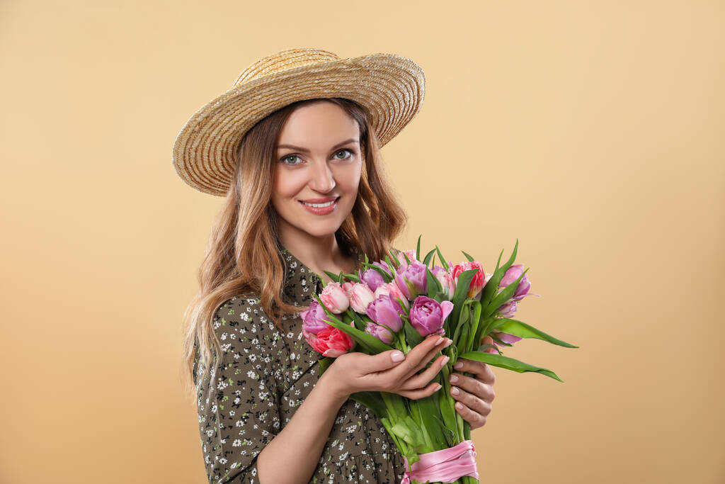 Щаслива молода жінка в солом'яному капелюсі тримає букет красивих тюльпанів на бежевому фоні
 - Фото, зображення