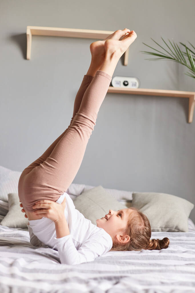 Cute little girl doing workout in bedroom at home on bad having fun raised her legs keeps hands in hips doing exercise birch in bedroom morning training and stretching. - Photo, Image