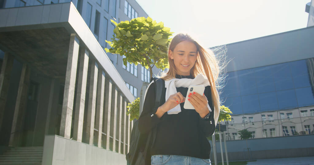 Гарний вигляд задоволена усміхненою молодою світло-волохатою дівчиною в повсякденному одязі з рюкзаком, що стоїть біля офісної будівлі і використовує свій мобільний в сонячний день
 - Фото, зображення