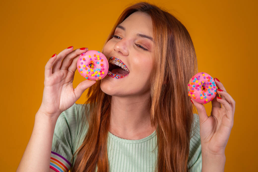 Menina de cabelos vermelhos no fundo amarelo comendo deliciosos donuts coloridos. - Foto, Imagem
