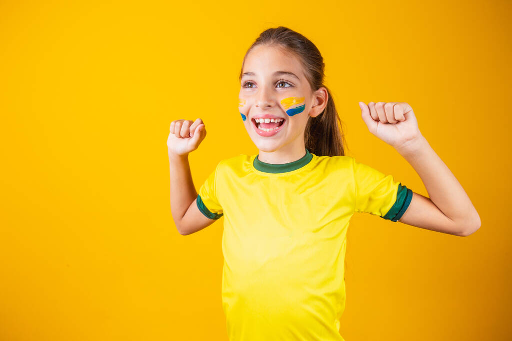Piękna dziewczynka kibicująca swojej drużynie na żółtym tle. Mała dziewczynka świętuje cel i świętuje zwycięstwo Brazylii - Zdjęcie, obraz