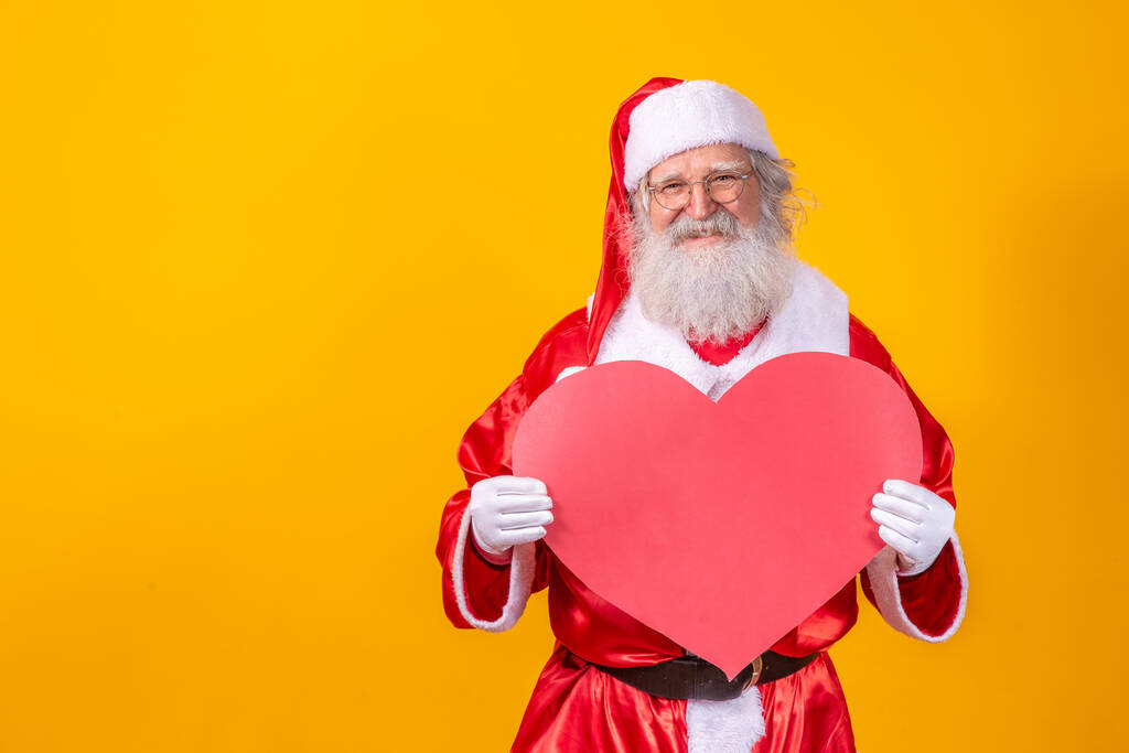Der Weihnachtsmann hält eine große rote Herzkarte vor gelbem Hintergrund. Romantischer Weihnachtsmann. - Foto, Bild