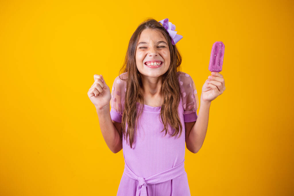 Щаслива світлошкіра дівчинка їсть фіолетове морозиво, приправлене виноградом, стискає кулак з очікуванням, що станеться щось дивовижне, має зубну посмішку, носить фіолетовий літній одяг
 - Фото, зображення
