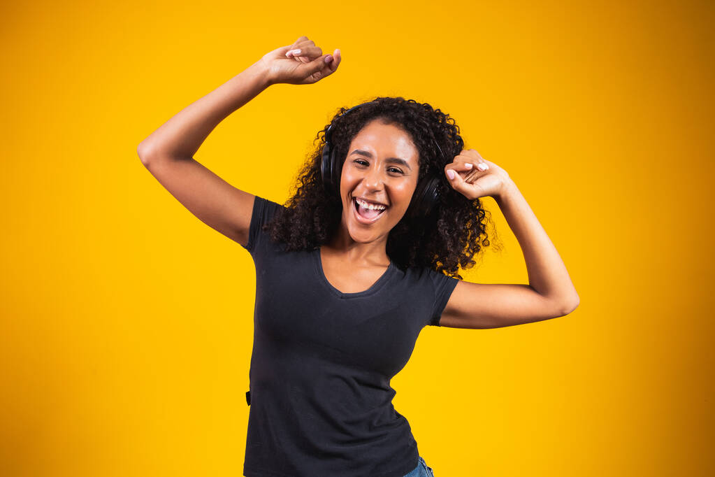 Jonge Afrikaanse vrouw draagt hoofdtelefoon dansend actief geïsoleerd op gele muur luisterend naar muziek lachend vrolijk. - Foto, afbeelding