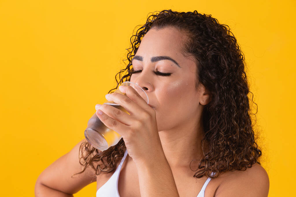 mujer afro joven bebiendo agua en fondo amarillo. Chica joven con vaso de agua. Una joven bebiendo agua del vaso. Concepto de hidratación - Foto, Imagen
