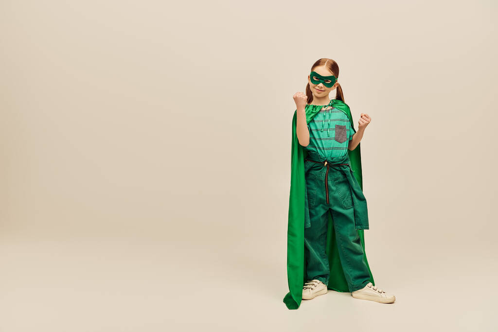 krachtig meisje in groene superheld kostuum met mantel en masker op het gezicht, het dragen van een broek en t-shirt en staan met gebalde vuisten, terwijl vieren Kinderbescherming dag vakantie op grijze achtergrond  - Foto, afbeelding