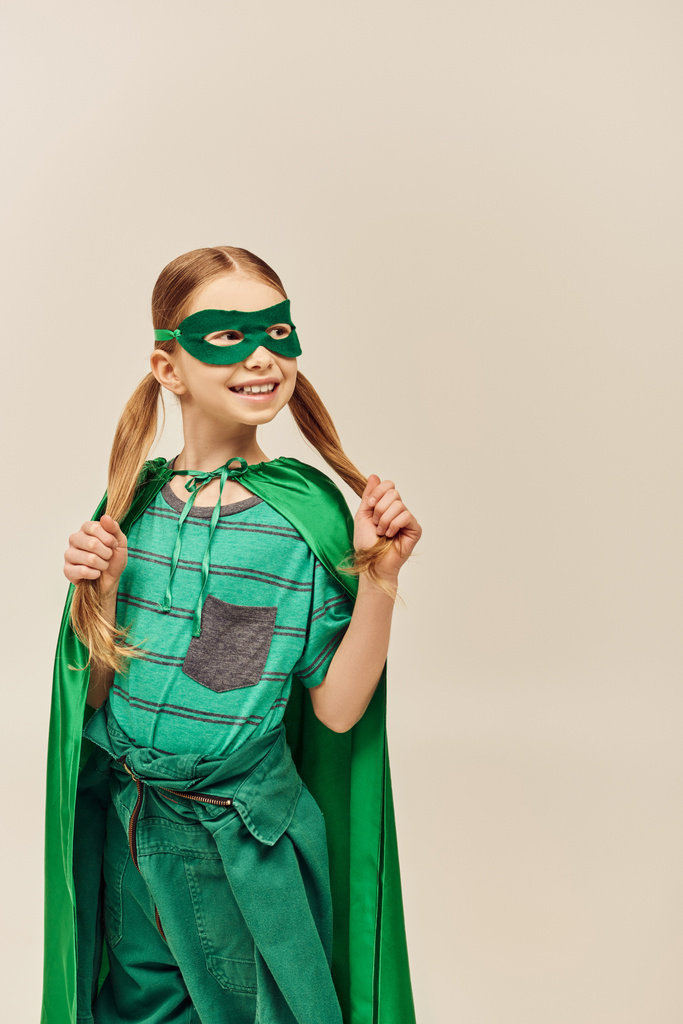 glückliches Mädchen im grünen Superheldenkostüm mit Mantel und Maske im Gesicht, mit Zwillingsschwanzfrisur, die ihr Haar berührt, während sie den Weltkinderschutztag auf grauem Hintergrund feiert  - Foto, Bild