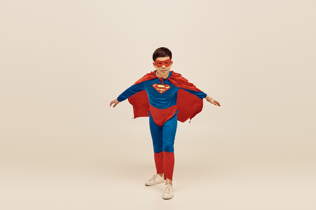 Mutiger asiatischer Junge im rot-blauen Superheldenkostüm mit Mantel und Maske im Gesicht anlässlich des Internationalen Tages zum Schutz von Kindern auf grauem Hintergrund  - Foto, Bild