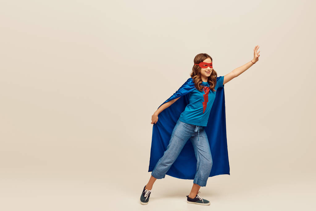 gelukkig meisje in superheld kostuum met blauwe mantel en rood masker op gezicht, staan in spijkerbroek en t-shirt met uitgestrekte hand, terwijl het bereiken van iets op grijze achtergrond  - Foto, afbeelding