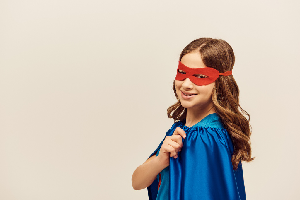 幸せな女の子でスーパーヒーロー衣装で青マントと赤マスク上の顔を見ながらカメラと笑顔で国際子供の日を祝います灰色の背景  - 写真・画像