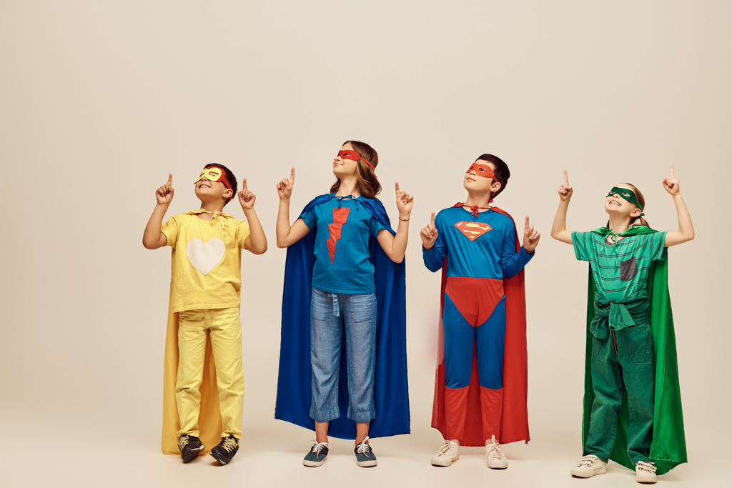positive multicultural kids in colorful superhero costumes with cloaks and masks pointing with fingers while celebrating Child protection day holiday on grey background in studio  - Photo, Image