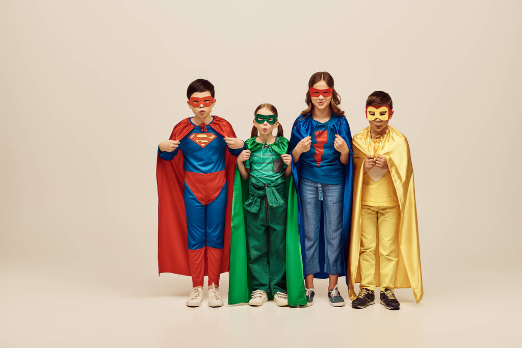 multicultural kids in colorful costumes with cloaks and masks pouting lips, looking at camera together and celebrating International children's day on grey background in studio  - Photo, Image