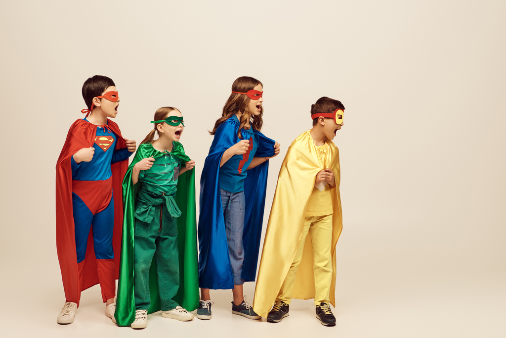 enfants multiculturels courageux en costumes colorés avec des manteaux et des masques criant et debout avec les poings serrés ensemble sur fond gris en studio, concept de Journée de la protection de l'enfance - Photo, image