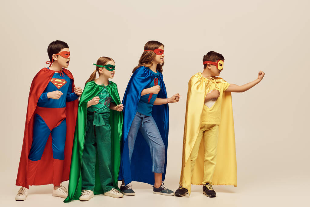 rohkeat monikulttuuriset lapset värikkäissä puvuissa, joissa on kaavut ja naamiot, jotka seisovat puristetut nyrkit yhdessä harmaalla taustalla studiossa, lastensuojelupäivän käsite - Valokuva, kuva