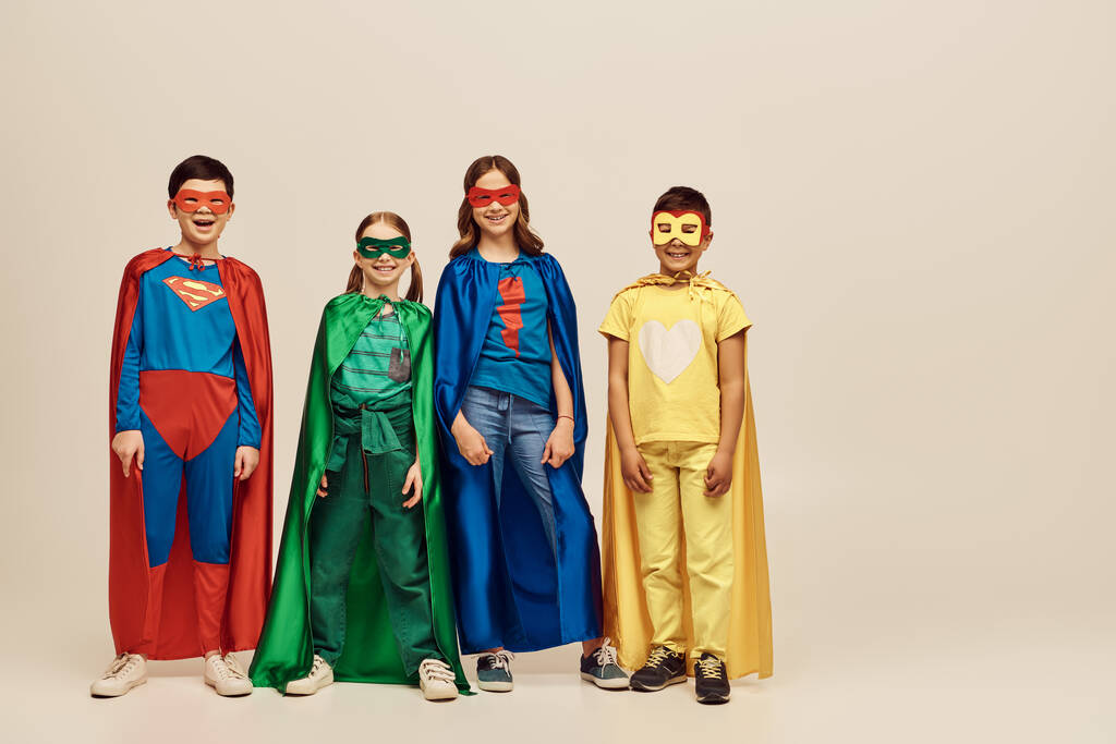 счастливые межрасовые дети в красочных костюмах в плащах и масках, улыбающиеся вместе и смотрящие в камеру на сером фоне в студии, концепция Дня защиты детей  - Фото, изображение