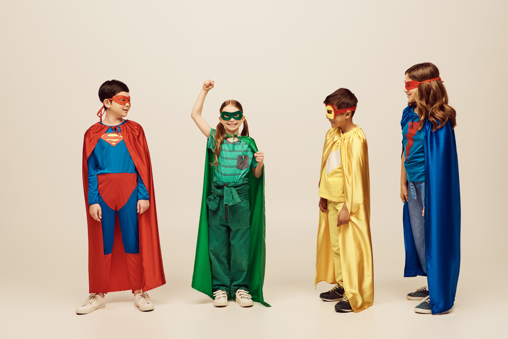 crianças inter-raciais felizes em trajes coloridos olhando para a menina em verde roupa de super-herói em pé com a mão levantada e protestando em fundo cinza no estúdio, conceito de Dia de Proteção à Criança  - Foto, Imagem