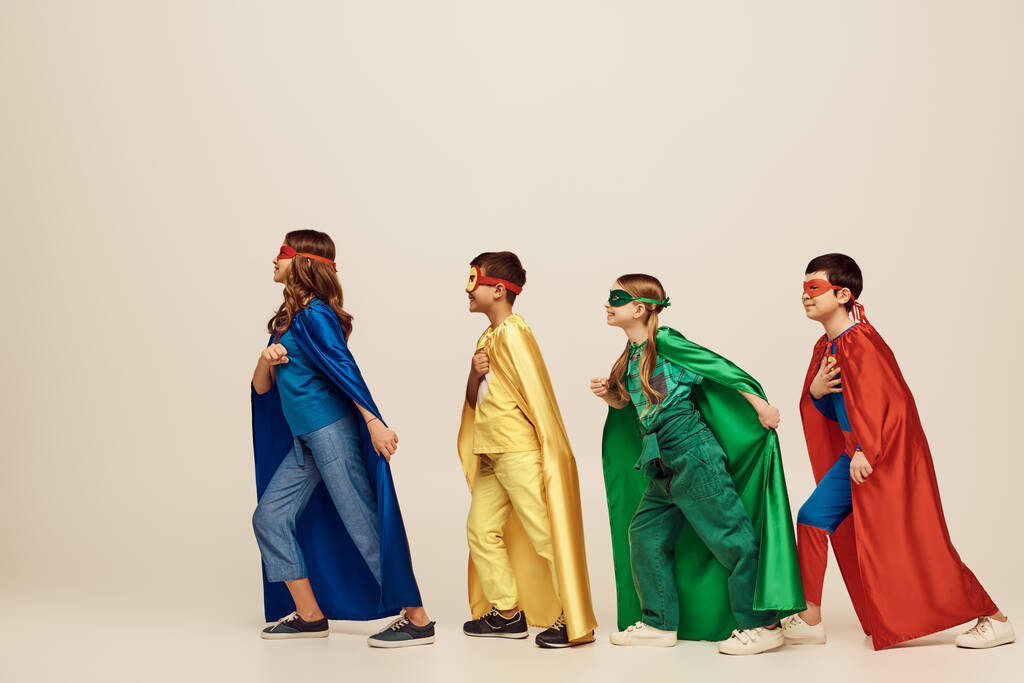 vista lateral de crianças inter-raciais felizes em trajes coloridos com capas e máscaras sorrindo e andando juntos em fundo cinza no estúdio, conceito de Dia de Proteção à Criança  - Foto, Imagem