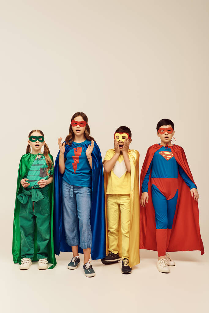 impactó a los niños interracial en trajes de superhéroes coloridos con capas y máscaras mirando a la cámara en el fondo gris en el estudio, concepto del Día de la Protección Infantil  - Foto, Imagen