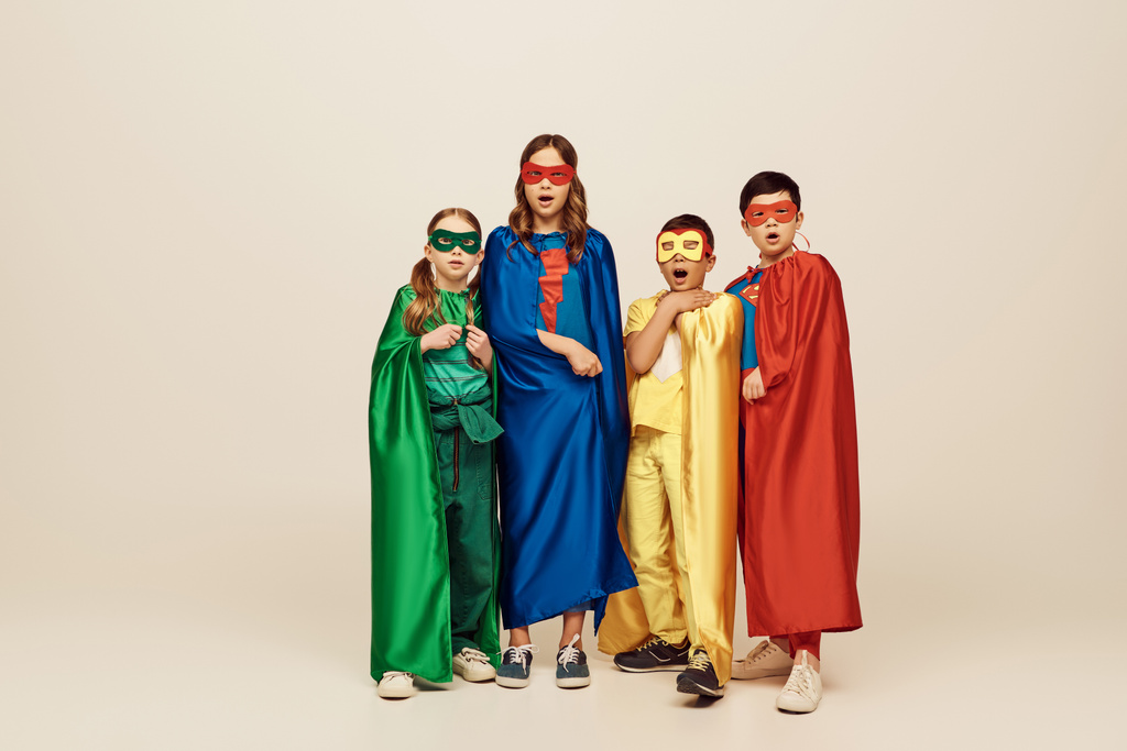 geschokt multiculturele en tiener kinderen in kleurrijke superheld kostuums met mantels en maskers kijken naar camera op grijze achtergrond in studio, Internationale kinderdag concept  - Foto, afbeelding
