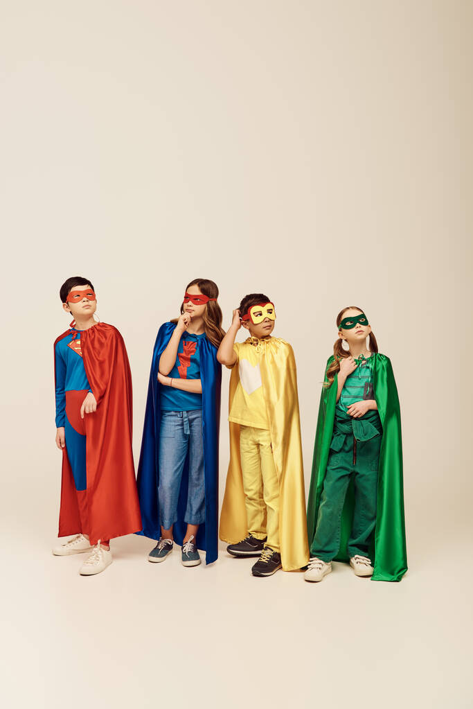 pensive multiculturele kinderen in kleurrijke superheld kostuums met mantels en maskers krabben hoofd en weg te kijken terwijl het denken over grijze achtergrond in de studio, kinderdag concept - Foto, afbeelding