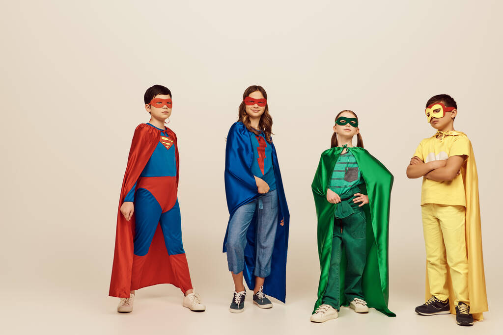 カラフルなスーパーヒーローの衣装を着た異人種間の子供たちマスクとクロークが立って、スタジオで灰色の背景に一緒にポーズ、国際子供の日のコンセプト - 写真・画像