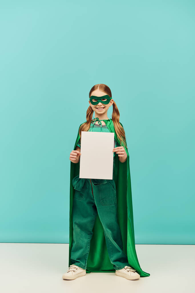 heureuse fille préadolescente en costume de super-héros vert avec manteau et masque debout avec du papier vierge et regardant la caméra tout en célébrant les vacances de la journée de protection de l'enfance sur fond bleu  - Photo, image