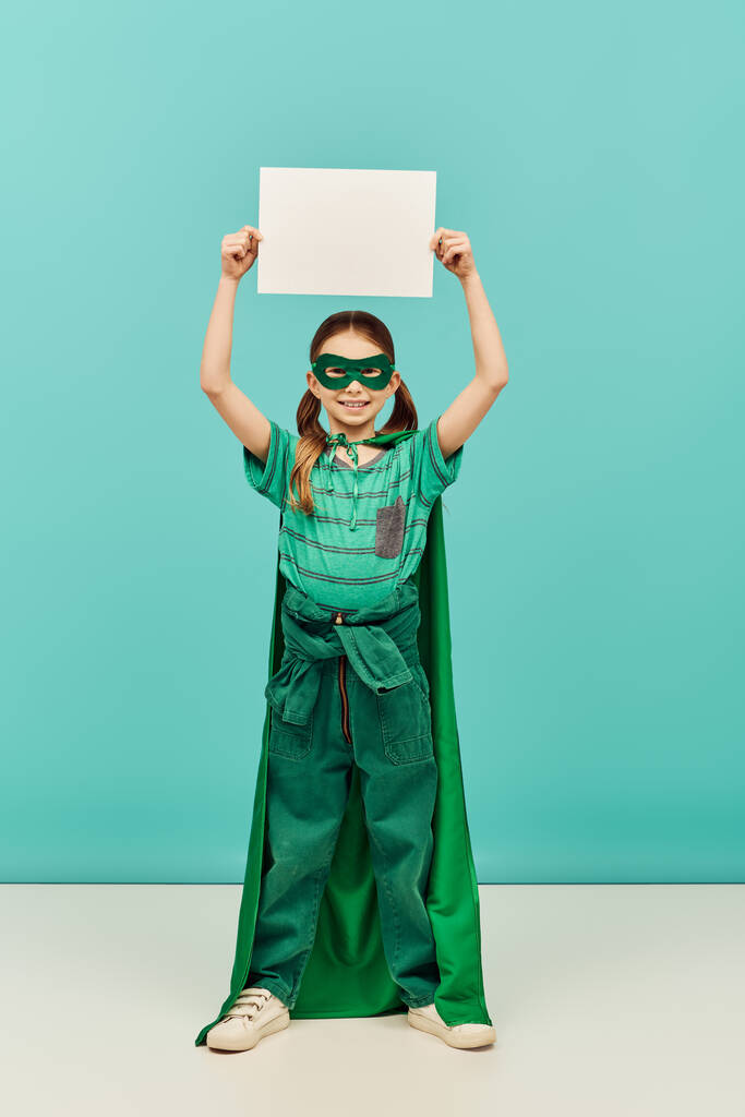 glückliches Mädchen im grünen Superheldenkostüm mit Mantel und Maske, das leeres Papier über dem Kopf hält und in die Kamera schaut, während es den Kinderschutztag auf blauem Hintergrund feiert  - Foto, Bild