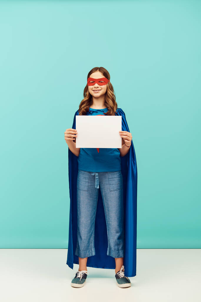 позитивная девочка-подросток в костюме супергероя с плащом и красной маской, стоящей с чистой бумагой и смотрящей в камеру во время празднования Дня защиты детей на синем фоне  - Фото, изображение