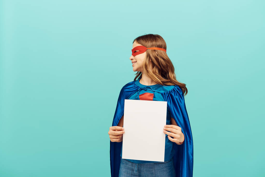 беззаботная девочка-подросток в костюме супергероя с красной маской, стоящей с чистой бумагой и глядя в сторону на синий фон, концепция Всемирного дня защиты детей  - Фото, изображение