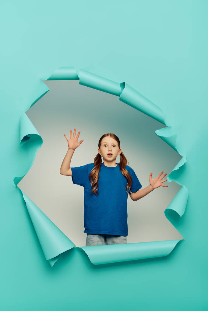 verängstigtes rothaariges Mädchen, das in die Kamera schaut, während es gestikuliert, in der Nähe eines blauen zerrissenen Papierlochs auf weißem Hintergrund, Konzept für den Internationalen Kindertag  - Foto, Bild
