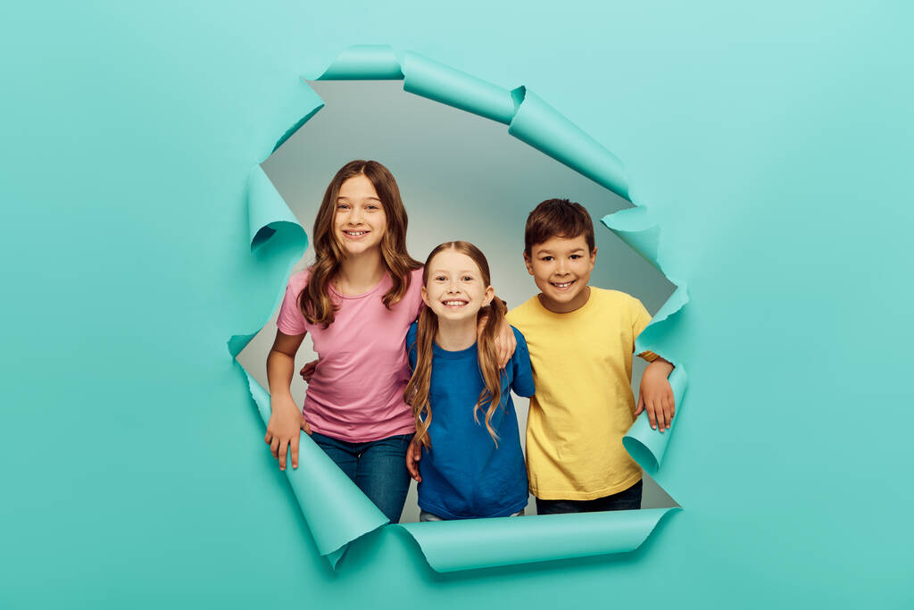 Positive interracial preteen kids in colorful t-shirts looking at camera while celebrating child protection day and standing behind hole in blue paper background - Photo, Image