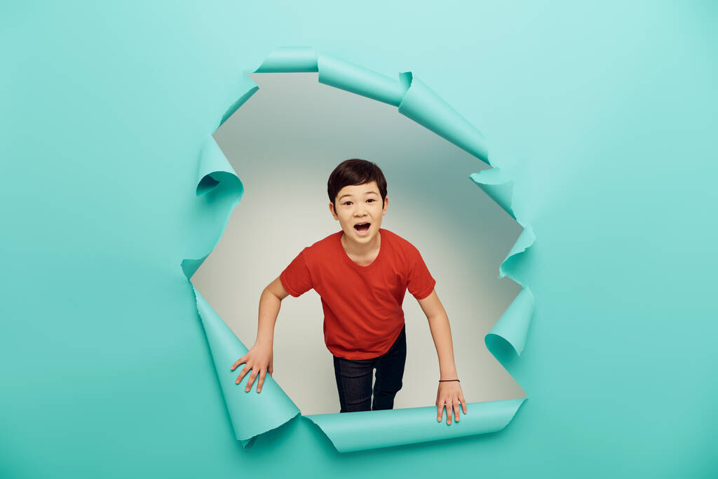 Astonished asian preteen kid in casual red t-shirt looking at camera during child protection day celebration behind hole in blue paper on white background - Photo, Image