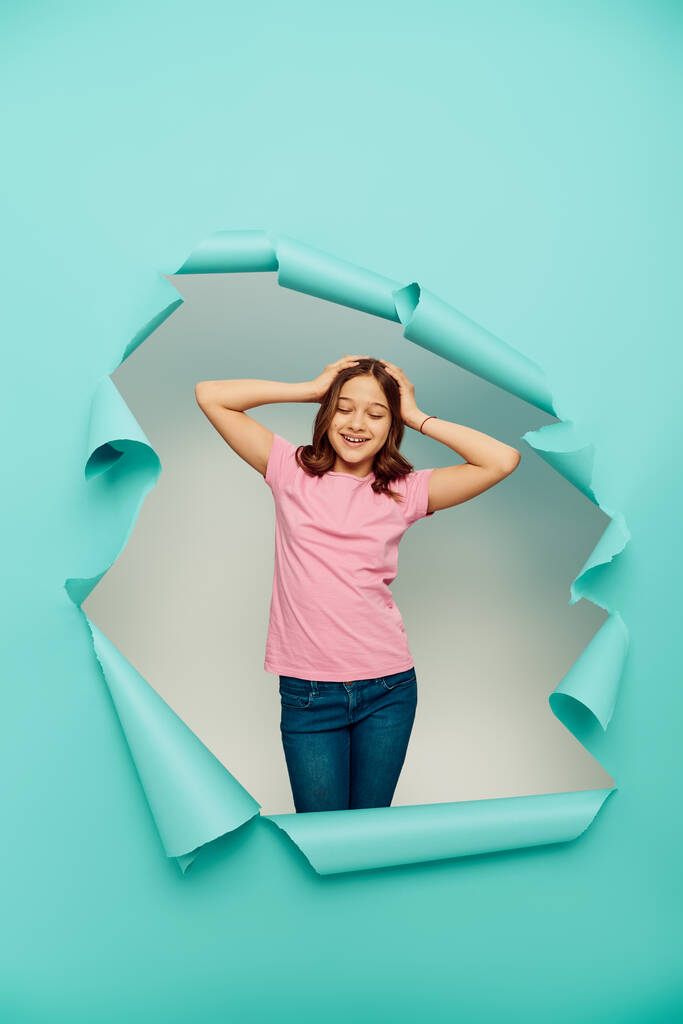 Positive preteen girl in pink t-shirt touching hair while standing behind hole in blue paper on white background, Happy children's day concept  - Photo, Image