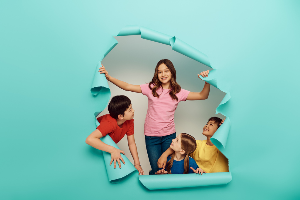 Smiling interracial kids in colorful clothes looking at friend while celebrating international children day behind hole in blue paper background - Photo, Image