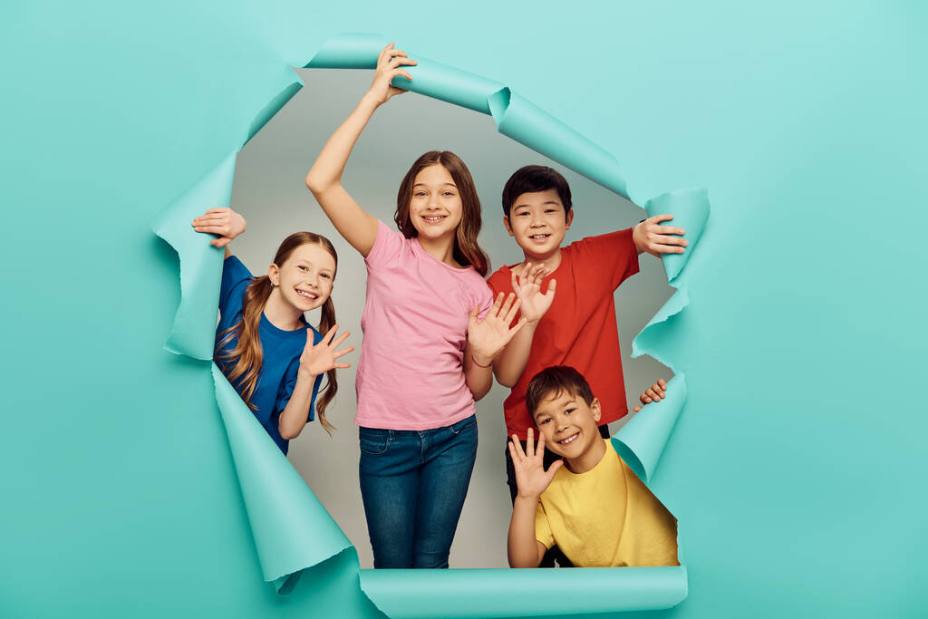 Smiling interracial kids in casual clothes waving hands at camera during international children day celebration behind hole in blue paper background - Photo, Image