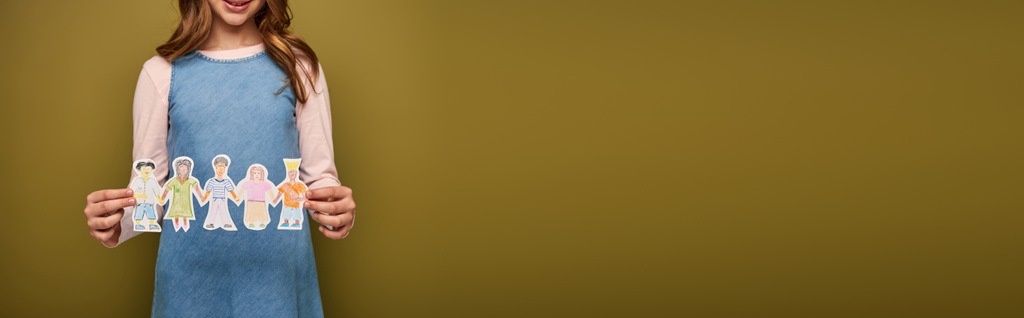 Vista recortada del niño preadolescente sonriente en vestido de mezclilla sosteniendo personajes de papel dibujado durante el día de los niños de protección mundial sobre fondo caqui, pancarta  - Foto, imagen