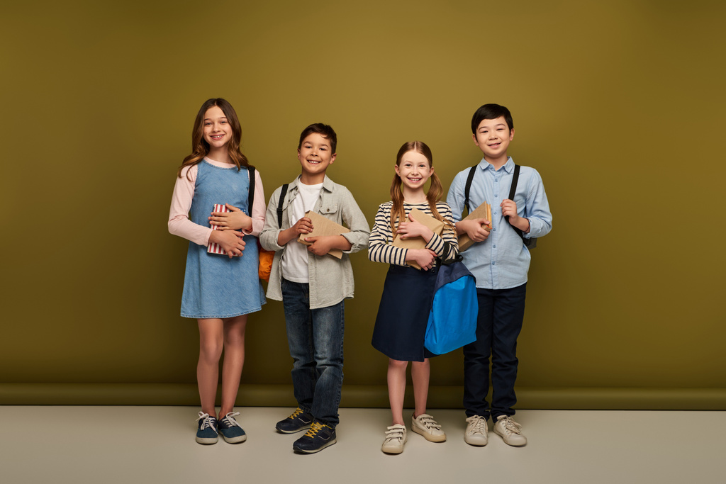 Full length of cheerful multiethnic preteen kids in casual clothes holding backpacks and books on khaki background, Happy children's day concept  - Photo, Image