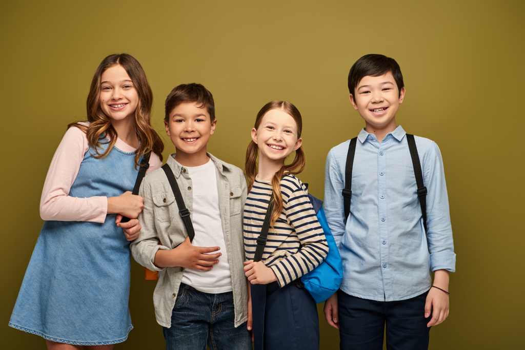 Mosolygó többnemzetiségű gyerekek hátizsák alkalmi ruhák nézi a kamerát, miközben együtt állnak a gyermekvédelmi nap ünneplés khaki háttér - Fotó, kép