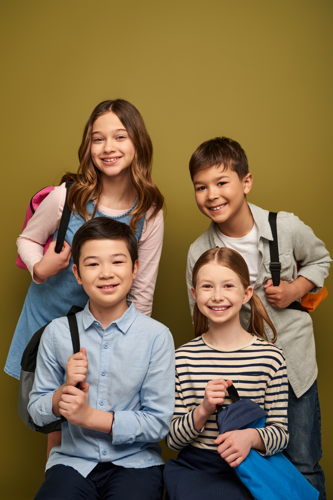 Enfants souriants multiethniques en vêtements décontractés tenant des sacs à dos et regardant la caméra pendant la célébration de la journée de protection de l'enfance sur fond kaki - Photo, image