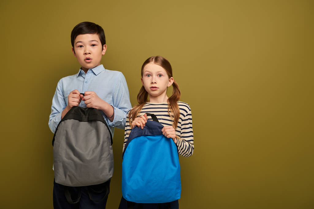 Τρομαγμένα πολυεθνικά προεφηβικά παιδιά με καθημερινά ρούχα κρατώντας σακίδια και κοιτάζοντας την κάμερα μαζί κατά τη διάρκεια του εορτασμού της διεθνούς ημέρας για την προστασία των παιδιών στο χακί φόντο - Φωτογραφία, εικόνα