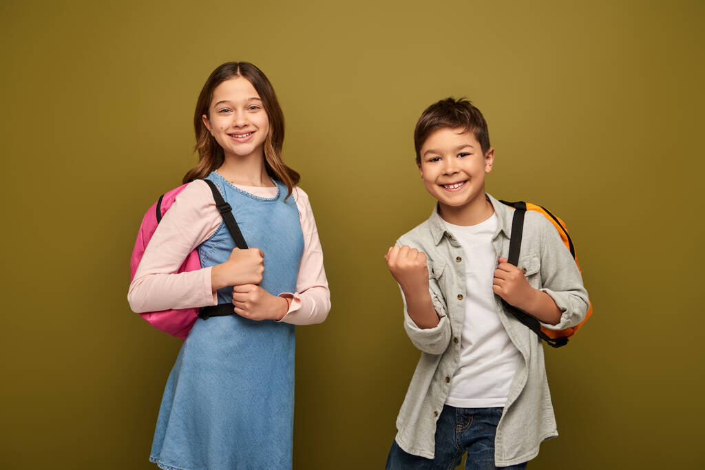 Excited multiracial boy with backpack showing yes gesture and looking at camera near friend in dress during child protection day celebration on khaki background - Photo, Image