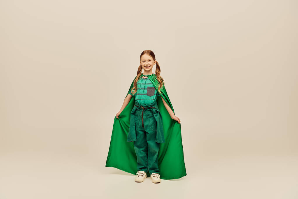 Veselý ryšavý kluk v zeleném kostýmu superhrdiny drží pláštěnku a dívá se na kameru, zatímco stojí na šedém pozadí ve studiu během mezinárodní dětské oslavy - Fotografie, Obrázek