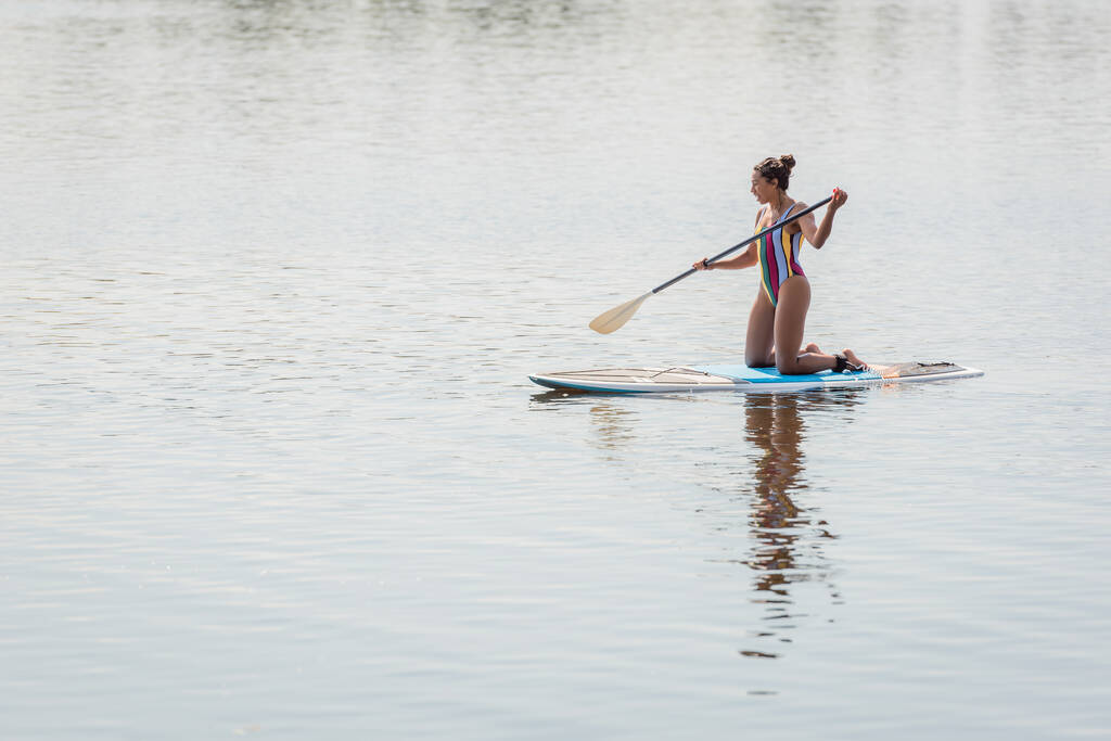 slender african american woman in colorful swimsuit holding paddle while standing on knees and sailing on sup board on river during weekend on summer day - Photo, Image