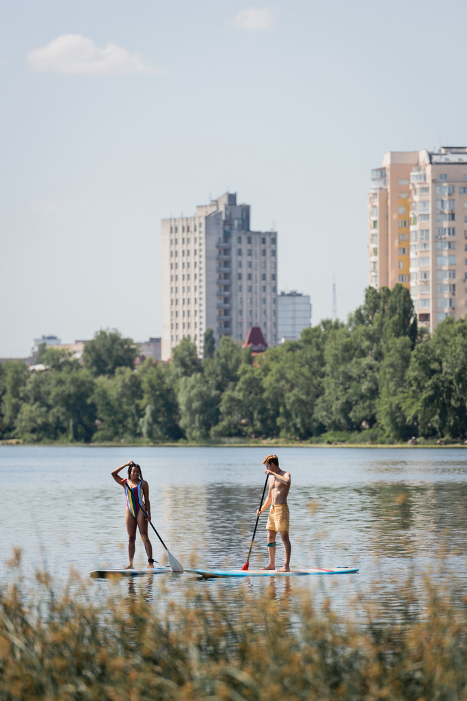 спортивна міжрасова пара в купальнику, що пливе на дошках з веслами, проводячи час на озері з мальовничим міським пейзажем і рослинами на розмитому передньому плані
 - Фото, зображення