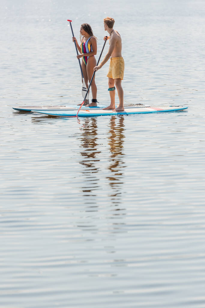 full length of young redhead man in swim shorts and brunette african american woman in colorful swimsuit sailing on sup boards on calm river on summer day - Photo, Image