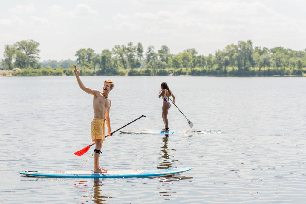 homme rousse joyeux regardant loin et pointant avec la main près sportive femme afro-américaine en maillot de bain coloré voile sur planche à soup le jour d'été avec bord de rivière pittoresque sur fond - Photo, image