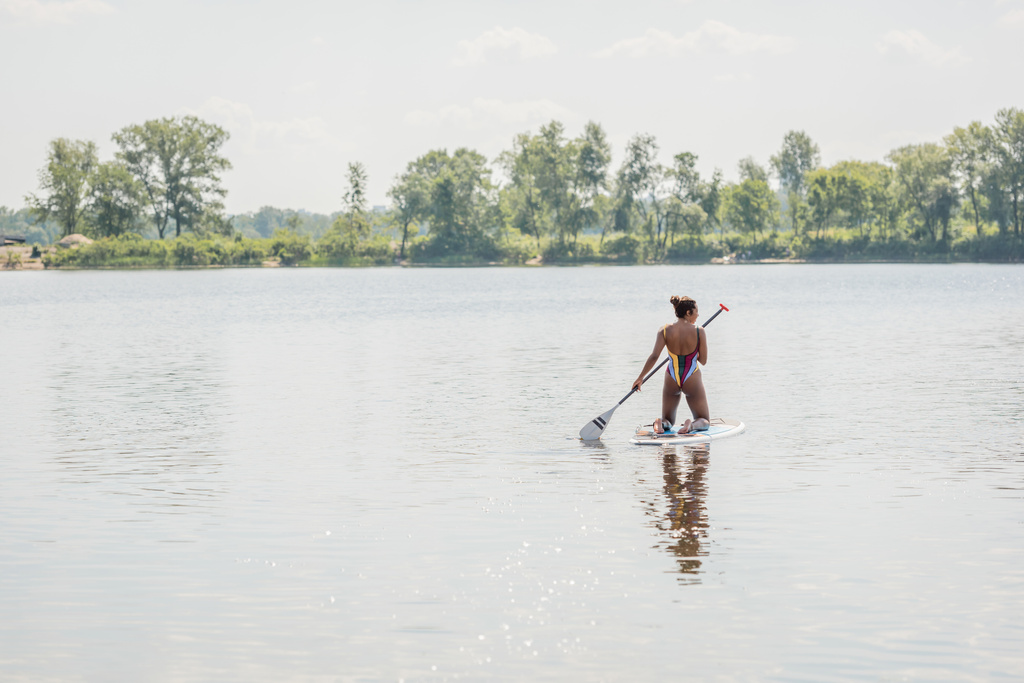 achteraanzicht van actieve Afro-Amerikaanse vrouw in gestreepte zwembroek met rust op de rivier door te zeilen op sup board in de buurt van rivier met groene bomen, outdoor activiteit, zomer vibes - Foto, afbeelding