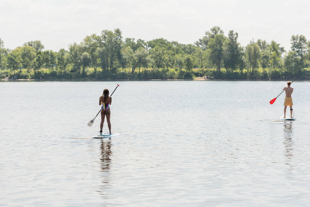 задний вид спортивного мужчины и африканской американки в красочных купальниках, плавающих на досках с веслами на озере с зеленым живописным берегом, активностью на открытом воздухе, летними вибрациями - Фото, изображение